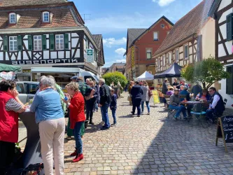 Eine belebte Straßenansicht mit Menschen, die an Ständen essen und trinken. Im Hintergrund sind typische Fachwerkhäuser und ein klarer blauer Himmel zu sehen.