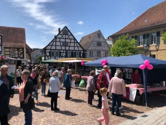 Ein belebter Wochenmarkt mit vielen Menschen in einer malerischen Stadt. Die Stände sind mit bunten Schirmen und Ballons geschmückt.