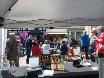 Ein lebhaftes Fest mit vielen Menschen in einer Marktsituation. Stände und Tische sind sichtbar, und die Atmosphäre ist fröhlich und gesellig.