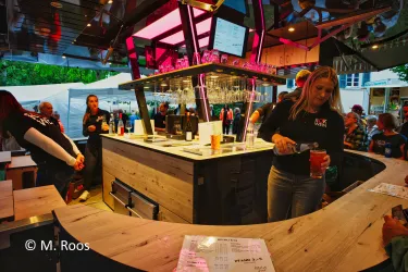 Eine moderne Bar mit einer Person, die ein Getränk einschenkt. Im Hintergrund sind Gäste und eine einladende Atmosphäre zu sehen.