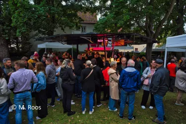 Eine lebhafte Veranstaltung im Freien mit einer großen Gruppe von Menschen, die um einen Stand versammelt sind. Bäume im Hintergrund und Zeltdächer schaffen eine einladende Atmosphäre.
