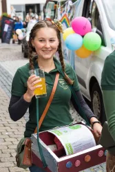 Ein fröhliches Mädchen mit Zöpfen hält ein Glas Getränk in der Hand. Sie trägt ein grünes T-Shirt und hat eine Kiste mit bunten Knöpfen vor sich.