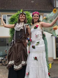 Zwei junge Frauen in fantasievollen Kostümen stehen fröhlich nebeneinander. Die eine trägt ein elegantes weißes Kleid mit Blumen, die andere ein braunes, gestreiftes Outfit mit einem Einhorn-Horn.
