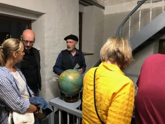 Eine Gruppe von Menschen steht in einem Raum und schaut auf einen Globus. Ein älterer Mann erklärt etwas, während die anderen interessiert zuhören.