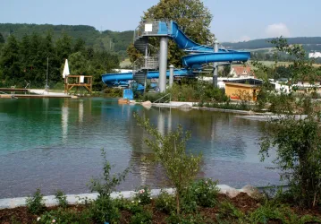 Ein Schwimmbad mit blauen Rutschen und klaren Wasserflächen. Im Hintergrund sind Bäume und eine grüne Landschaft zu sehen.