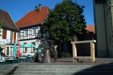 Ein schöner Platz mit einer Bronzeskulptur und einem großen Baum. Im Hintergrund sind traditionelle Gebäude und Sitzgelegenheiten im Freien sichtbar.