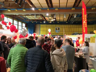Eine lebendige Messe mit vielen Menschen, die miteinander sprechen und inmitten von bunten Luftballons stehen. Im Hintergrund sind mehrere Informationsstände und Banner zu sehen.