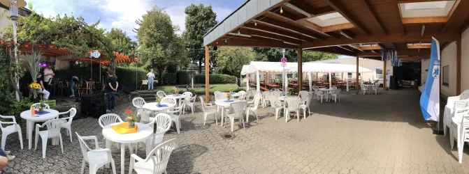 Ein Außenbereich mit weißen Tischen und Stühlen in einem Garten. Im Hintergrund sind Bäume und eine überdachte Terrasse zu sehen.