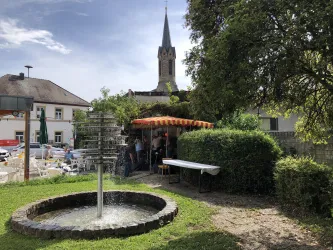 Ein gemütlicher Biergarten mit einem Brunnen inmitten von grünen Büschen. Im Hintergrund ist der Kirchturm sichtbar, und einige Gäste genießen die frische Luft.