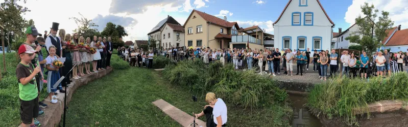 Eine große Menschenmenge versammelt sich an einem Wasserlauf, um eine Veranstaltung zu beobachten. Im Hintergrund sind mehrere Häuser und ein klarer Himmel zu sehen.