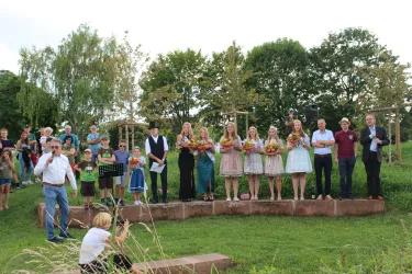 Eine festliche Zeremonie im Freien mit mehreren Menschen auf einer Bühne. Die meisten stehen in traditionellen Kleidern und halten Blumensträuße.