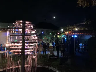 Eine entspannte Abendatmosphäre mit einer beleuchteten Wasserfontäne und Menschen, die sich im Garten aufhalten. Der Mond scheint am Nachthimmel.