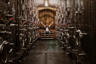 Ein Weinlager mit metallenen Tanks und einem Mann, der stolz in der Mitte sitzt. Die Atmosphäre ist rustikal und einladend.