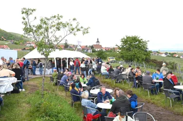 Eine lebhafte Veranstaltung im Freien mit vielen Menschen, die an Tischen sitzen und sich unterhalten. Im Hintergrund sind Zelte und eine malerische Landschaft zu sehen.