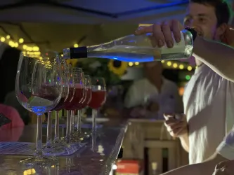 Ein Barkeeper gießt Wein in Gläser an einer Bar. Im Hintergrund sind Lichter und Menschen zu sehen.