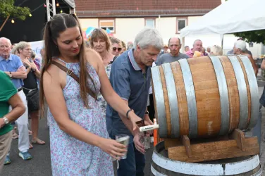 Eine Frau zapft ein Getränk aus einem kleinen Holzfass auf einem festlichen Ereignis. Im Hintergrund sind zahlreiche Menschen zu sehen, die die Veranstaltung genießen.