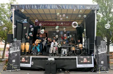 Eine Band spielt auf einer Bühne, umgeben von vielen Seifenblasen. Im Hintergrund sind Bäume und ein Banner mit dem Text „Jungwinzer Weinfest“ zu sehen.