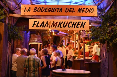 Eine belebte Markthalle mit mehreren Menschen, die sich um Stände versammeln. Im Vordergrund sind Schilder mit den Beschriftungen "La Bodeguita del Medio" und "Flammkuchen" zu sehen.