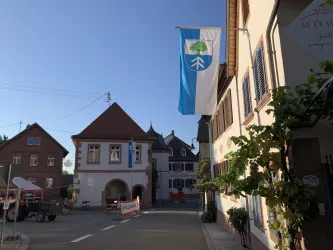 Ein ruhiger Dorfplatz mit alten Gebäuden und einer Fahne, die ein Baum-Symbol zeigt. Der Himmel ist blau und wolkenlos.
