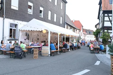 Eine belebte Straßenszene mit einem Zelt, in dem Menschen an Tischen sitzen und essen. Die Umgebung zeigt traditionelle Gebäude und eine gemütliche Atmosphäre.