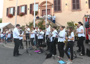 Eine Blaskapelle tritt auf einer Straße in einer kleinen Stadt auf. Im Hintergrund stehen Zuschauer und genießen die Musik.