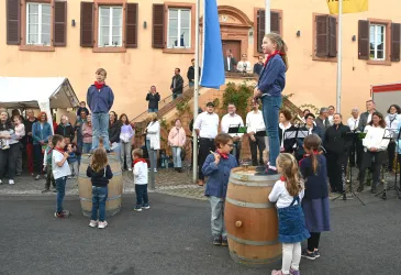 Eine Gruppe von Kindern steht auf großen Fässern und feiert eine Veranstaltung im Freien. Im Hintergrund sind viele Zuschauer und traditionelle Gebäude zu sehen.