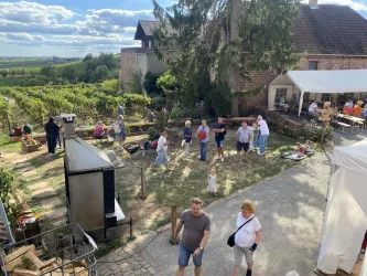 Eine lebhafte Veranstaltung im Freien mit Menschen, die sich in einem Garten aufhalten. Im Hintergrund sind Weinreben und ein festliches Zelt zu sehen.