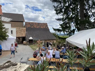 Eine gemütliche Outdoor-Veranstaltung mit vielen Besuchern an langen Tischen. Im Hintergrund sind Gebäude und Bäume zu sehen, die eine ländliche Atmosphäre schaffen.