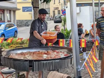 Ein spanischer Koch steht hinter einer großen Pfanne und bereitet spanische Paella zu. Die Szene findet auf einem öffentlichen Platz statt.