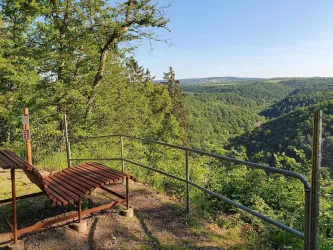 Ein Aussichtspunkt mit einer Holzliege und Blick auf die bewaldete Landschaft. Die Sonne scheint auf einen klaren Himmel, was eine ruhige Atmosphäre schafft.