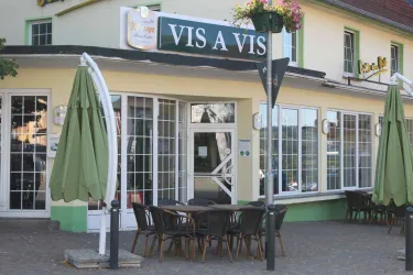 Ein Restaurant mit dem Namen "VIS A VIS", das große Fenster und eine Außenterrasse mit Tischen und Stühlen hat. Die Markisen sind grün und verleihen dem Ambiente eine einladende Atmosphäre.