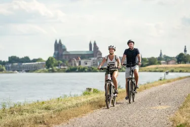Zwei Personen fahren mit dem Fahrrad entlang eines Flusses. Im Hintergrund sind historische Gebäude und eine schöne Landschaft zu sehen.