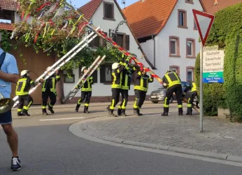 Eine Gruppe von Feuerwehrleuten trägt einen festlich geschmückten Baum durch eine Straße. Im Hintergrund sind typische deutsche Häuser und eine Verkehrsschilder zu sehen.