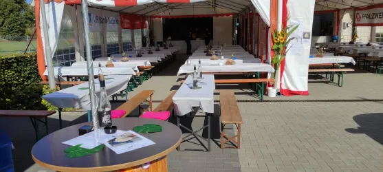 Ein festlich dekoriertes Zelt mit langen Tischen und Bänken. Die Tische sind mit weißen Tischdecken gedeckt und der Raum ist gut beleuchtet.