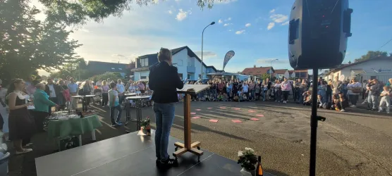 Eine Person steht auf einer Bühne und spricht zu einer großen Menschenmenge. Im Hintergrund sind Gebäude und ein klarer Himmel zu sehen.