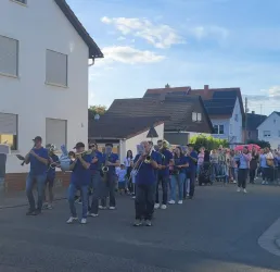 Eine Blaskapelle spielt Straßenmusik, während eine Menschenmenge entlang der Häuser zieht. Die Veranstaltung findet an einem klaren Tag statt.