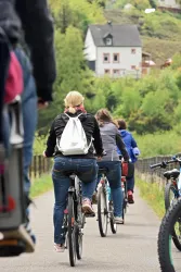 Eine Gruppe von Radfahrern bewegt sich auf einem Radweg. Im Hintergrund sind grüne Hügel und ein weißes Haus zu sehen.
