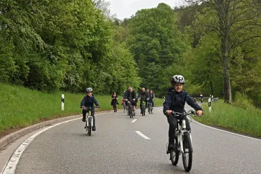 Eine Gruppe von Kindern fährt mit Fahrrädern auf einer ruhigen Straße umgeben von Bäumen. Der Tag ist bewölkt und die Natur wirkt grün und einladend.