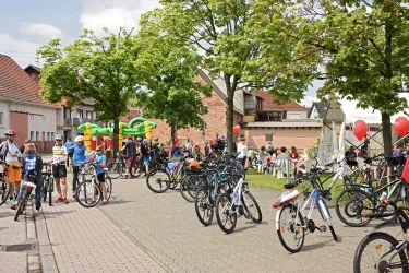Eine belebte Straße mit vielen Fahrrädern und Menschen. Im Hintergrund sind grüne Bäume und ein Spielbereich für Kinder zu sehen.