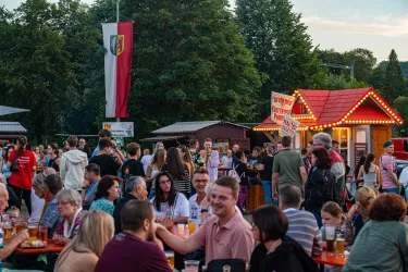 Eine lebhafte Menschenmenge bei einem Fest im Freien. Stände mit Beleuchtung und viele Tische mit Getränken sind zu sehen.