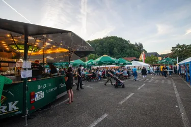 Ein belebtes Festival mit zahlreichen Ständen und Besuchern. Im Hintergrund sind grüne Hügel und ein klarer Himmel zu sehen.