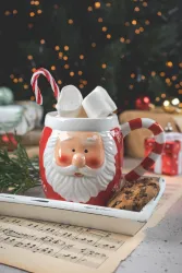 Eine festliche Tasse mit einem Weihnachtsmann-Motiv, gefüllt mit Marshmallows und einem Zuckerstängel. Daneben liegen Plätzchen und Weihnachtsdekoration auf dem Tisch.