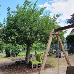Ein Spielplatz mit einer Schaukel und einem großen Baum. Im Hintergrund sind Personen mit Gartenarbeiten beschäftigt.