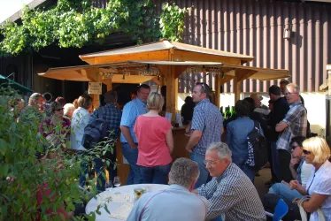 Eine lebhafte Veranstaltung im Freien mit vielen Menschen, die sich um eine Holzbar versammeln. Die Atmosphäre ist entspannt und freundlich, umgeben von Pflanzen und einem rustikalen Gebäude.