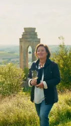 Eine Frau steht lächelnd auf einer Wiese mit einer Aussicht auf eine alte Ruine im Hintergrund. Die Landschaft ist grün und friedlich, was eine angenehme Atmosphäre schafft.