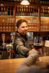 Ein Barkeeper reicht einem Gast ein Glas Rotwein an der Bar. Im Hintergrund sind zahlreiche Weinflaschen und Gläser sichtbar.