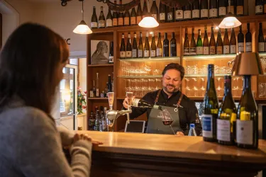 Ein Barkeeper serviert Wein an eine Kundin an der Theke. Im Hintergrund sind zahlreiche Weinflaschen und Gläser zu sehen.