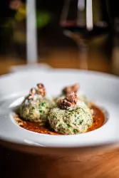 Zwei grüne Knödel mit Kräutern auf einer roten Sauce, garniert mit Nüssen. Im Hintergrund sind Weingläser zu sehen.