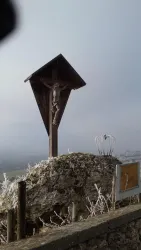 Ein Holz-Kreuz mit einer Christusfigur steht auf einem Felsen. Im Hintergrund sind neblige Himmel und frostige Landschaften zu sehen.