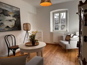 Ein helles Wohnzimmer mit Holzboden und großen Fenstern. Es gibt einen runden Tisch, einen Sessel und ein Kunstwerk an der Wand.